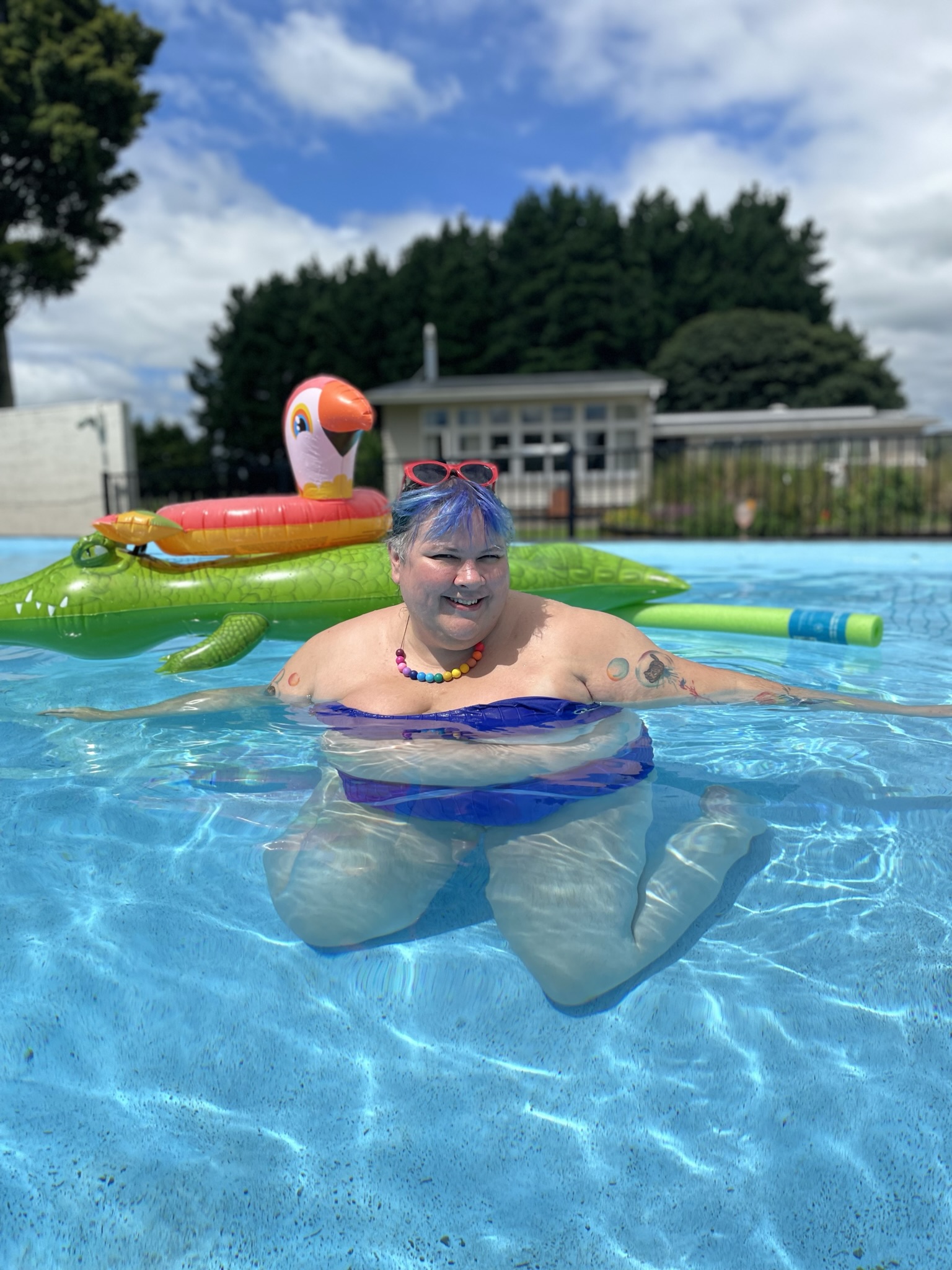 Joanna, a fat babe, wears a bikini in a glorious blue pool, pulled off her shoulders to show off a rainbow bead necklace. In the background are two